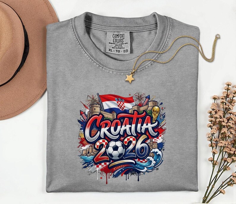 a gray t-shirt with a graphic design depicting the word "Croatia" surrounded by various elements such as a soccer ball, a castle, and a flag. The design also includes the text "Croatia 2026" and a star. The t-shirt is laid out on a white surface, with a hat and some flowers visible in the background.