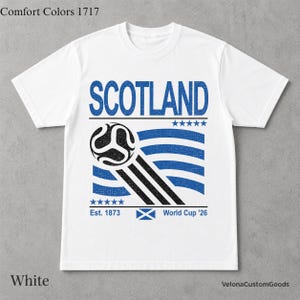 May include: A white short-sleeve t-shirt featuring a blue graphic with the text SCOTLAND, Est. 1873, and World Cup '26. The design includes a black and white soccer ball, blue wavy lines, ten blue stars, and a small blue and white Scottish flag.