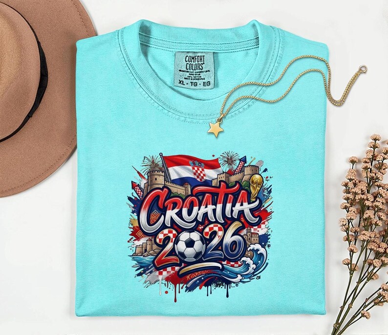 A turquoise t-shirt with a graphic design featuring the text "Croatia 2026" surrounded by various elements such as a soccer ball, flags, and buildings.