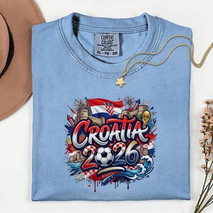 A blue t-shirt with a graphic design featuring the text "Croatia 2026" surrounded by various elements such as a soccer ball, a castle, and a star.