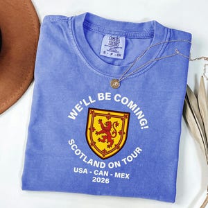 A blue t-shirt with a graphic design and text, along with a brown hat and some dried leaves, are arranged on a white surface.