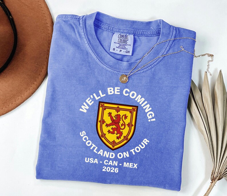 A blue t-shirt with a graphic design and text, along with a brown hat and some dried leaves, are arranged on a white surface.