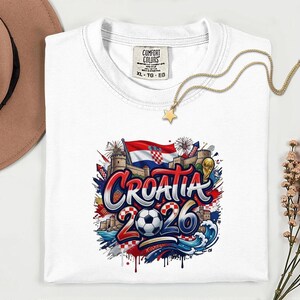 a white t-shirt with a graphic design depicting the word "Croatia" surrounded by various elements such as a soccer ball, flags, and buildings. The design also includes the text "Croatia 2026" and a star. The t-shirt is placed on a white surface, with a hat and some flowers visible in the background.