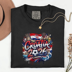 a black t-shirt with a graphic design depicting the word "Croatia" surrounded by various elements such as a soccer ball, flags, and buildings. The design also includes the text "Croatia 2026" and a star. The t-shirt is placed on a white surface, with a hat and some flowers visible in the background.