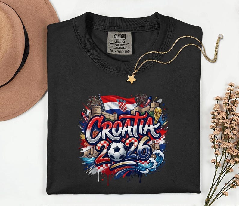 a black t-shirt with a graphic design depicting the word "Croatia" surrounded by various elements such as a soccer ball, flags, and buildings. The design also includes the text "Croatia 2026" and a star. The t-shirt is placed on a white surface, with a hat and some flowers visible in the background.