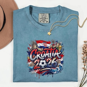 a blue t-shirt with a graphic design depicting the word "Croatia" surrounded by various elements such as a soccer ball, flags, and a castle. The t-shirt is laid out on a white surface, with a hat and some flowers nearby.