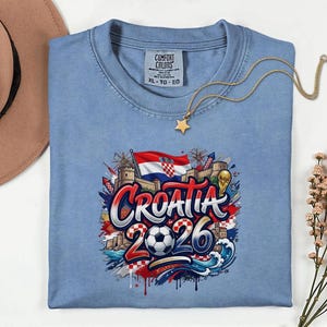 a blue t-shirt with a graphic design depicting the word "Croatia" surrounded by various elements such as a soccer ball, a castle, and a flag. The t-shirt is laid out on a white surface, with a hat and some flowers nearby.