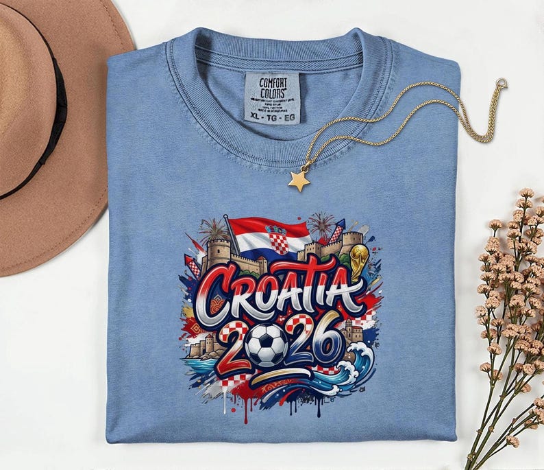 a blue t-shirt with a graphic design depicting the word "Croatia" surrounded by various elements such as a soccer ball, a castle, and a flag. The t-shirt is laid out on a white surface, with a hat and some flowers nearby.