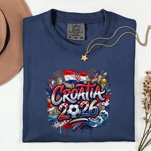 a blue t-shirt with a graphic design depicting the word "Croatia" surrounded by various elements associated with the country, such as a soccer ball, a castle, and a star. The t-shirt is laid out on a white surface, with a hat and some flowers nearby.
