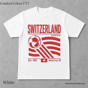 May include: A white short-sleeve t-shirt featuring a red graphic print. The design includes the text SWITZERLAND, a soccer ball, five stars, the text Est. 1895, a Swiss flag icon, and the text World Cup '26. The text Comfort Colors 1717 is visible.