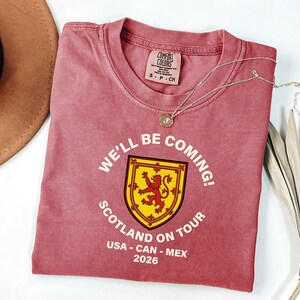 A red t-shirt with a yellow and red shield design, along with the text "WE'LL BE COMING! SCOTLAND ON TOUR USA - CAN - MEX 2026", is displayed on a white surface alongside a brown hat and some leaves.