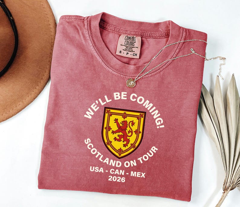 A red t-shirt with a yellow and red shield design, along with the text "WE'LL BE COMING! SCOTLAND ON TOUR USA - CAN - MEX 2026", is displayed on a white surface alongside a brown hat and some leaves.
