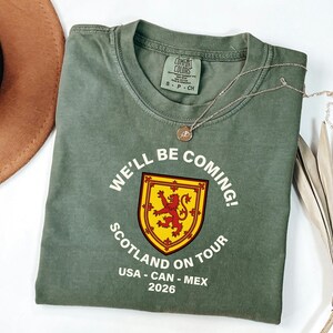 a green t-shirt with a red and yellow shield design and text, along with a brown hat and some dried leaves, all laid out on a white surface.