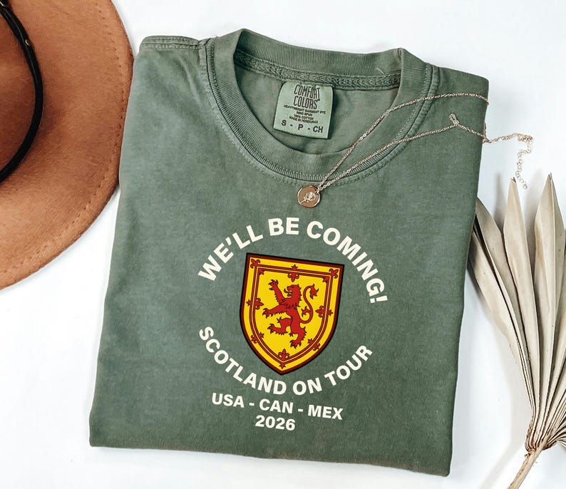 a green t-shirt with a red and yellow shield design and text, along with a brown hat and some dried leaves, all laid out on a white surface.