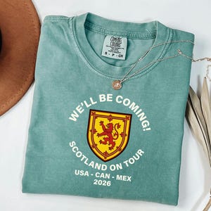 A light blue t-shirt with a graphic design and text, along with a brown hat and some dried leaves, are arranged on a white surface.