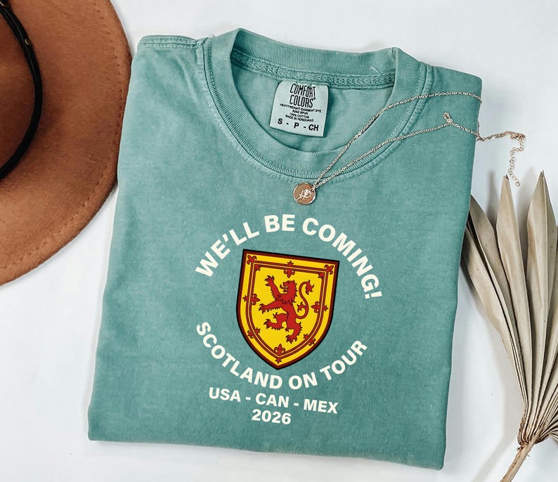 A light blue t-shirt with a graphic design and text, along with a brown hat and some dried leaves, are arranged on a white surface.