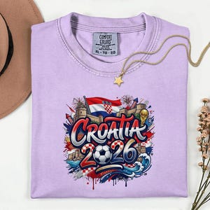 a purple t-shirt with a graphic design depicting the word "Croatia" surrounded by various elements such as a soccer ball, a castle, and a star. The t-shirt is placed on a white surface, with a hat and some flowers nearby.