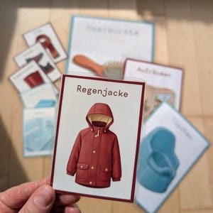 Könnte beinhalten: Eine rechteckige laminierte Karte mit einer zentrierten Illustration einer dunkelroten Regenjacke mit Kapuze, cremefarbenem Innenfutter, Knöpfen auf der Vorderseite und zwei Pattentaschen. Das Wort Regenjacke steht in dunkelbrauner Schrift darüber.
