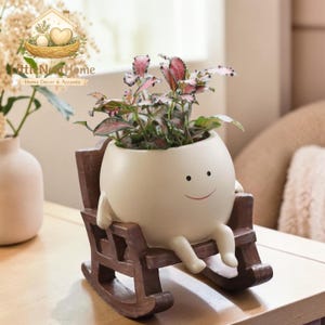 May include: A quirky, cream-coloured ceramic planter in the shape of a smiling character, seated in a rocking chair. The planter contains a small plant with pink and green leaves. The rocking chair is dark brown. Home decor and accents.