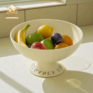 Op de afbeelding: Een crèmekleurige fruitschaal op voetstuk gevuld met een verscheidenheid aan vers fruit, waaronder een banaan, citroenen, pruimen en appels. De kom heeft een textuur aan de buitenkant en de woorden "FRUICE LIFE" staan op de basis.