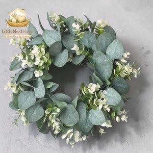 May include: A circular wreath featuring soft green eucalyptus leaves and small white flowers. The wreath is a home decor accent, perfect for adding a touch of nature to any space. The logo "LittleNestHome" is in the top left corner.