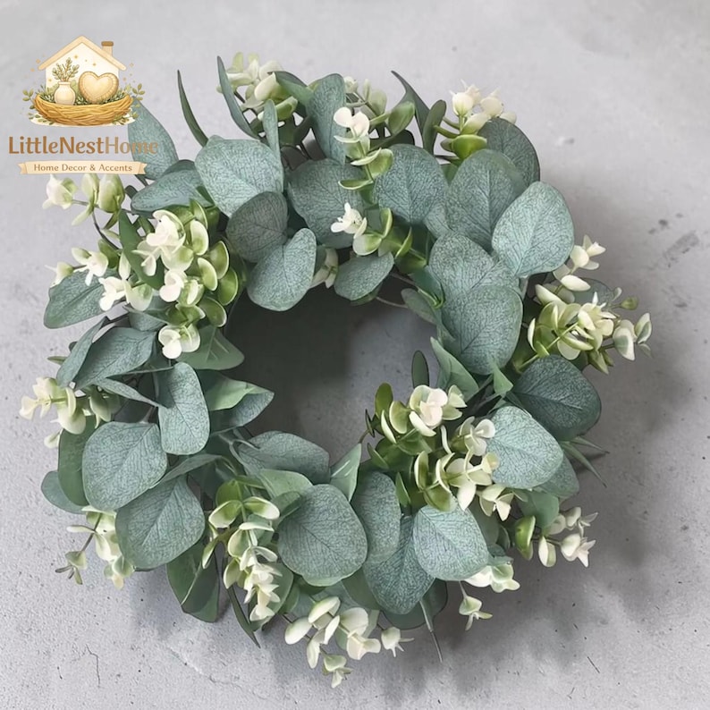 May include: A circular wreath featuring soft green eucalyptus leaves and small white flowers. The wreath is a home decor accent, perfect for adding a touch of nature to any space. The logo "LittleNestHome" is in the top left corner.