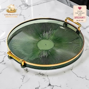 May include: A circular, green glass tray with a ribbed pattern, gold-coloured metal handles and trim. The tray sits on a white marble surface. The text "Click the heart for discount!" is visible. The tray is a decorative item.