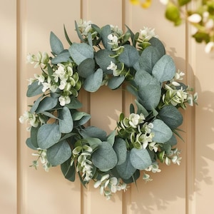 May include: A circular wreath composed of large, muted green eucalyptus leaves and small white flowers. The wreath is a decorative item, suitable for hanging on a door or wall. The leaves have a soft, textured appearance.
