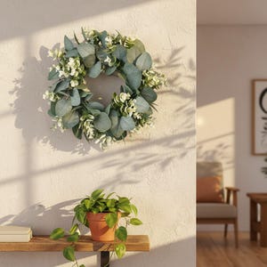 May include: A decorative wreath featuring green eucalyptus leaves and small white flowers. The wreath is hanging on a textured, off-white wall. A small potted plant with green leaves sits on a wooden shelf below.
