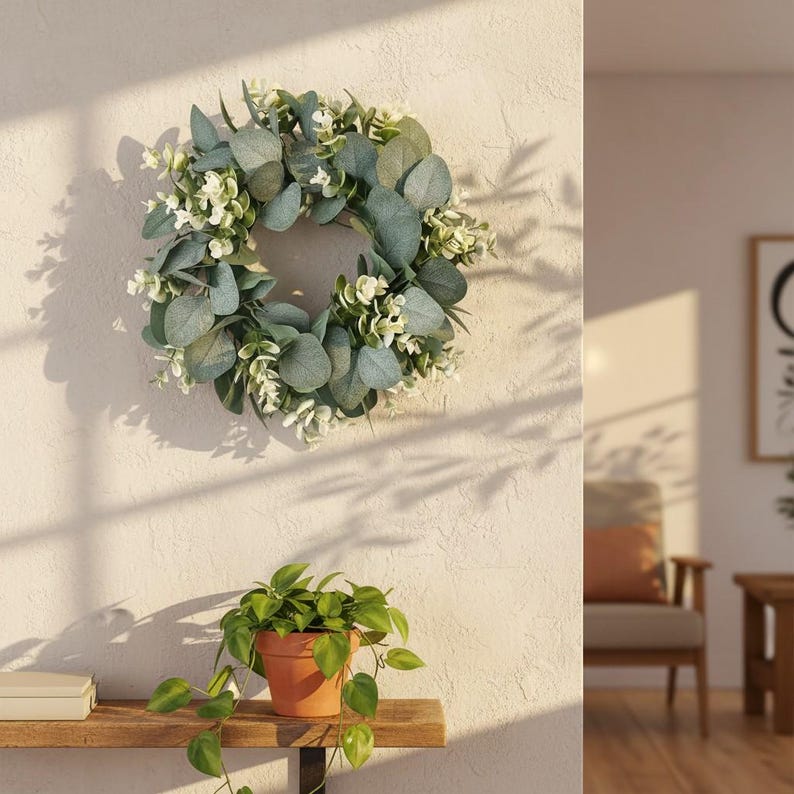 May include: A decorative wreath featuring green eucalyptus leaves and small white flowers. The wreath is hanging on a textured, off-white wall. A small potted plant with green leaves sits on a wooden shelf below.