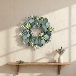 May include: A circular wreath featuring blue-green eucalyptus leaves and small white flowers. The wreath is displayed on a beige wall, above a wooden shelf. A small white vase with dried flowers sits on the shelf.