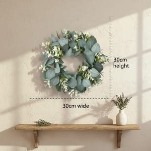 May include: A decorative wreath, 30cm wide and 30cm high, made of green eucalyptus leaves and white flowers. The wreath is hanging on a beige wall above a wooden shelf. A small white vase with dried plants is on the shelf.