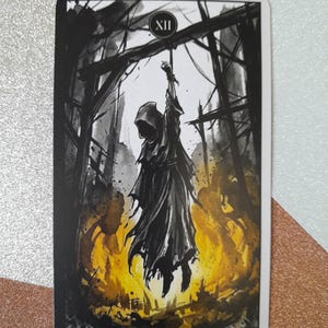 May include: A rectangular tarot card featuring a black and white ink-style illustration of a hooded figure suspended by a rope from a wooden structure. The figure is surrounded by vibrant yellow and orange flames at the base. The Roman numeral XII is at the top, and the text Hanged Woman is printed at the bottom.