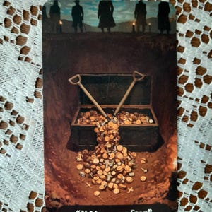 K&ouml;nnte beinhalten: Illustrierte Tarotkarte mit einer Schatzkiste, die mit Goldm&uuml;nzen &uuml;berquillt, und zwei Schaufeln. Silhouetten stehen auf einem H&uuml;gel im Hintergrund. Der Text "X Marks the Spot" steht unten.