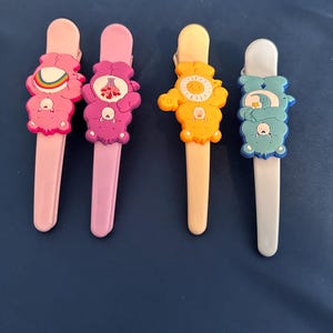 May include: Four colorful hair clips featuring cartoon bear designs. The clips are in shades of pink, purple, yellow, and blue. Each clip has a different bear design with a unique symbol on its belly.