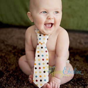 May include: A baby boy wearing a white and brown polka dot tie, sitting on a brown shag rug. The baby is smiling and looking to the right.