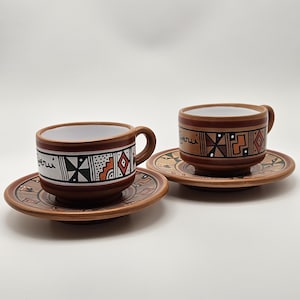 Può includere: Due tazze da caffè e piattini in ceramica con un design marrone e bianco. Le tazze e i piattini hanno un bordo marrone e un interno bianco. Le tazze hanno un manico. I piattini e le tazze hanno un motivo geometrico e la parola "Peru" è scritta sulle tazze.