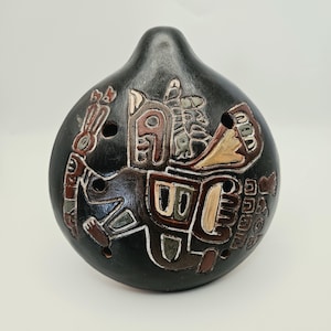Puede incluir: Una ocarina de cerámica negra de forma redonda con un diseño pintado a mano de una figura estilizada. El instrumento tiene varios orificios para los dedos dispuestos en su superficie, con tonos tierra en marrón, crema y verde apagado.