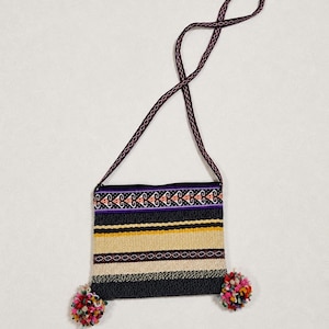 Può includere: Borsa a tracolla tessuta con un motivo a righe in nero, beige, giallo e grigio. La parte superiore presenta una fascia decorativa con disegni geometrici in rosa, viola e nero. Due pompon colorati pendono dal basso.