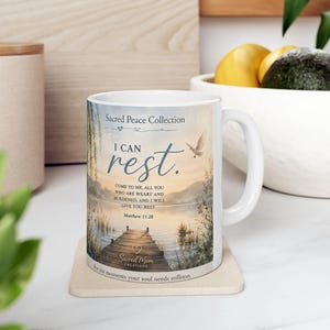 May include: A white ceramic mug featuring a printed landscape of a wooden dock extending into a calm lake at sunset. The text reads Sacred Peace Collection, I CAN rest., Matthew 11:28, and Sacred Mom Creations, with a flying white dove illustration.