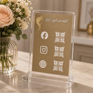 Könnte beinhalten: Klarer Acrylständer mit goldenem Hintergrund, der Social-Media-Symbole für Facebook, Instagram und eine Website sowie QR-Codes anzeigt. Auf dem Schild steht "let's get social" in weißer Schrift. Der Ständer steht auf einer transparenten Acrylbasis.