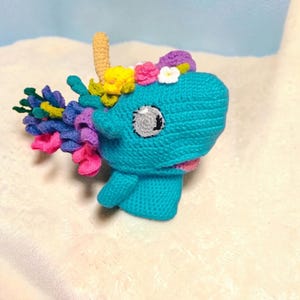 Könnte beinhalten: Ein handgefertigter, türkisfarbener Häkel-Einhornkopf mit einem beigen Horn, einem bunten Blumenkranz und einem rosa Mund. Das Einhorn hat ein graues Auge und eine fließende Mähne aus rosa, lila, blauer und grüner Wolle.