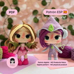 Puede incluir: Dos muñecas de ganchillo con grandes ojos negros y mejillas sonrosadas. La muñeca izquierda tiene pelo castaño, un conjunto rosa y alas verdes. La muñeca derecha tiene pelo morado, un gorro puntiagudo morado con un detalle verde y un traje morado.