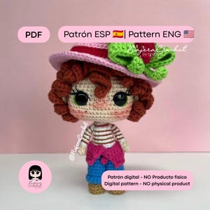 Puede incluir: Una muñeca de ganchillo con pelo rizado castaño rojizo, lleva un sombrero rosa con un lazo verde y un detalle de baya roja, una camiseta de rayas blancas y rojas, un suéter rosa atado a la cintura y pantalones azules. Texto: PDF, Patrón ESP, Pattern ENG.