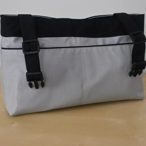 May include: A rectangular messenger bag with a light gray body and a black top section. The bag has black adjustable straps with plastic buckles. A thin black line accents the top of the gray section.