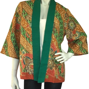 May include: A patterned, open-front jacket with a green collar and trim. The jacket features a brown and green batik design with floral and leaf motifs. The sleeves are three-quarter length. The jacket is displayed on a mannequin.