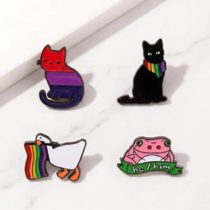 Puede incluir: Cuatro pines de esmalte con diseños de animales. Incluye un gato con rayas rojas, moradas y azules, un gato negro con una bufanda arcoíris, un ganso blanco con una bandera arcoíris y una rana rosa con el texto "he/him".