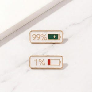 May include: Two rectangular enamel pins with gold borders. The top pin shows a battery at 99% with a green fill and a lightning bolt symbol. The bottom pin shows a battery at 1% with a red fill.