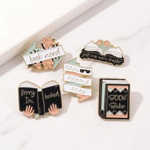 May include: A collection of enamel pins with book-themed designs. The pins feature phrases like "book nerd," "Cool People Read," and "Book Babe." The color palette includes black, white, pink, and mint green.