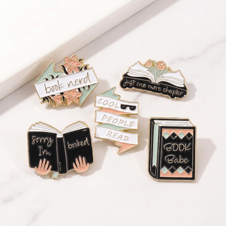 May include: A collection of enamel pins with book-themed designs. The pins feature phrases like "book nerd," "Cool People Read," and "Book Babe." The color palette includes black, white, pink, and mint green.
